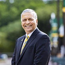 Ajay Vinze, dean of the School of Business, is standing outside on Wilkins Plaza. He is wearing a dark sportcoat, white button down shirt and a green and gold striped tie.